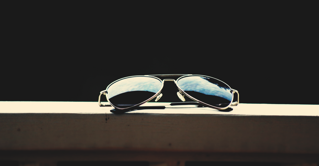 A close-up of aviator sunglasses reflecting the sky, set against a dark background.