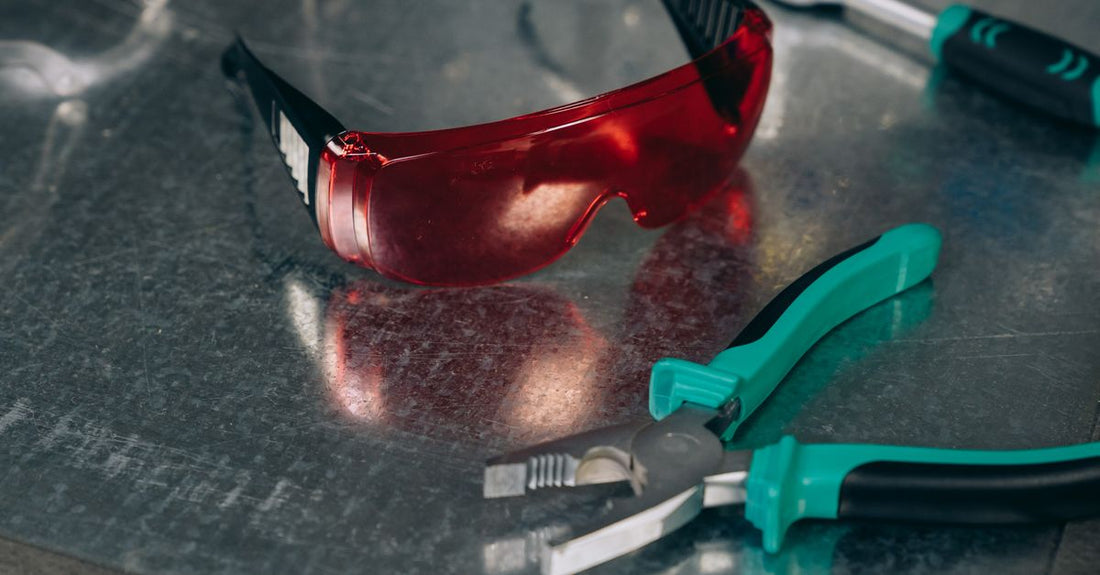 Red safety goggles and green-handled pliers on a reflective metal surface, emphasizing protective tools.